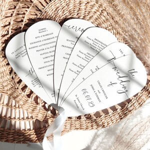 May include: A collection of white wedding ceremony fans with black calligraphy text, held together with a white ribbon. The fans are arranged in a woven basket. The text includes names, dates, and ceremony details.
