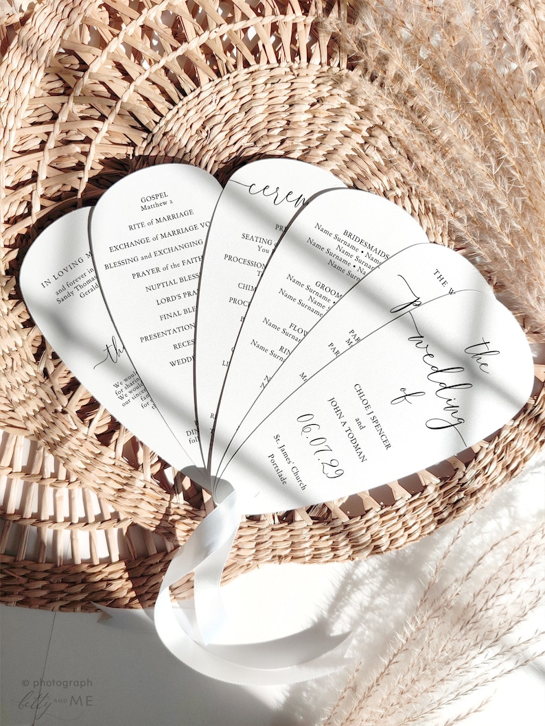 May include: A collection of white wedding ceremony fans with black calligraphy text, held together with a white ribbon. The fans are arranged in a woven basket. The text includes names, dates, and ceremony details.