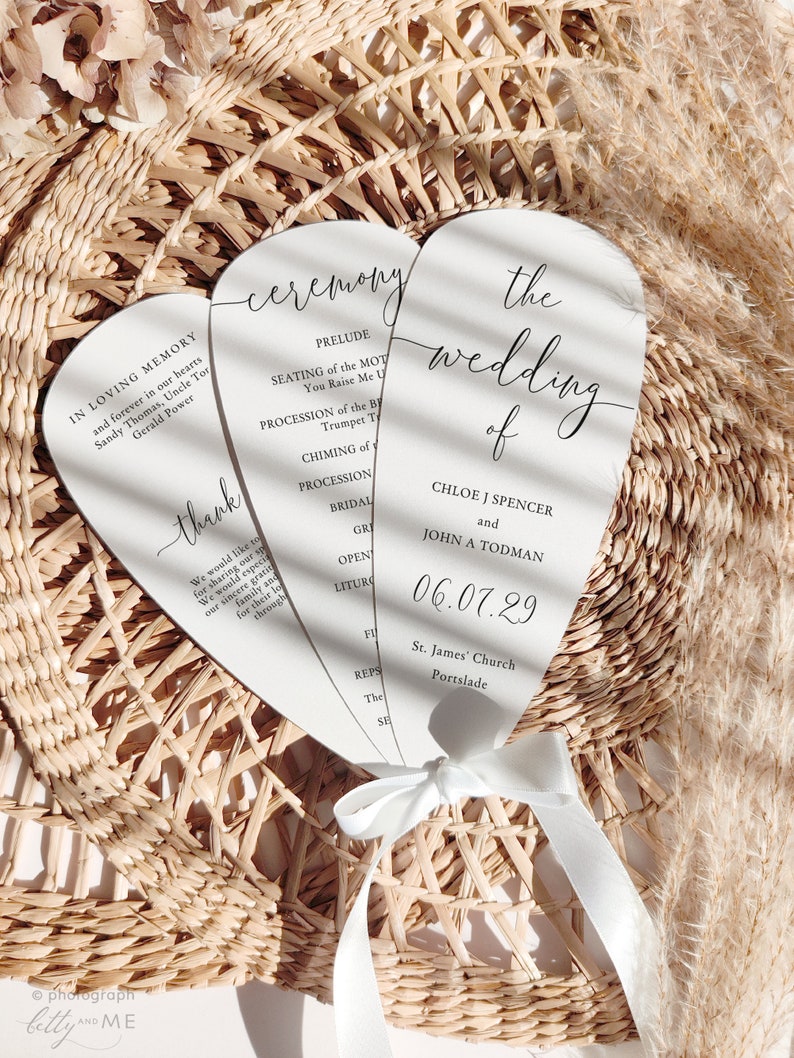 May include: Three white wedding ceremony programs tied with a white ribbon. The programs have black text with the words "ceremony" and "the wedding of" visible. The date "06.07.29" is also printed on the program.