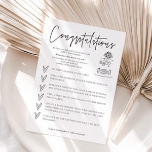 May include: A white card with the word "Congratulations" in script. The card lists tasks for a table captain, such as keeping the table lively and taking a selfie. Decorative icons of a tree, bottles, and cutlery are included.
