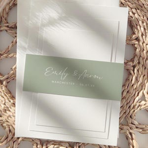 May include: A stack of white cards with a sage green belly band. The band features the names "Emily & Aaron" in elegant script, along with "Manchester - 06.07.XX". The background is a woven texture.