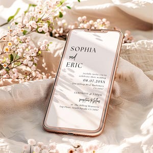 May include: A rose gold smartphone displays a wedding invitation. The invitation reads "SOPHIA and ERIC" with the date "08.07.20XX" and ceremony details. Soft pink flowers and a cream-colored fabric are in the background.