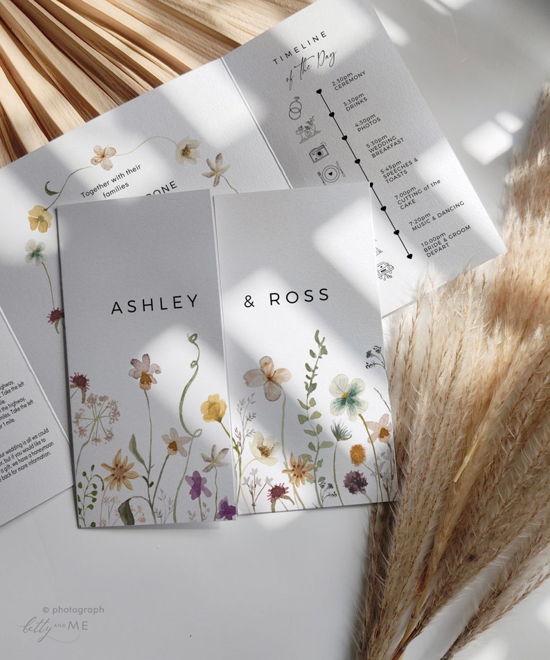 May include: A wedding invitation with a floral design. The invitation features a watercolor illustration of wildflowers in shades of purple, pink, yellow, and green. The text "Ashley & Ross" is printed in black at the top of the invitation. The invitation also includes a timeline of the wedding day.