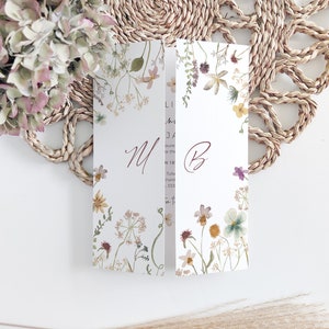 May include: A folded wedding invitation with floral watercolor illustrations. The invitation features the initials "M" and "B" in a burgundy script. The design includes a variety of flowers and greenery in soft pastel colors. The invitation is set against a woven placemat.