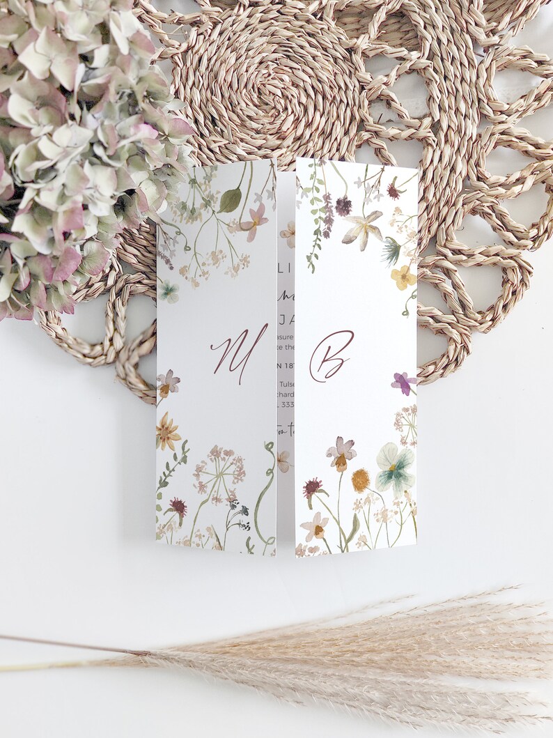 May include: A folded wedding invitation with floral watercolor illustrations. The invitation features the initials "M" and "B" in a burgundy script. The design includes a variety of flowers and greenery in soft pastel colors. The invitation is set against a woven placemat.
