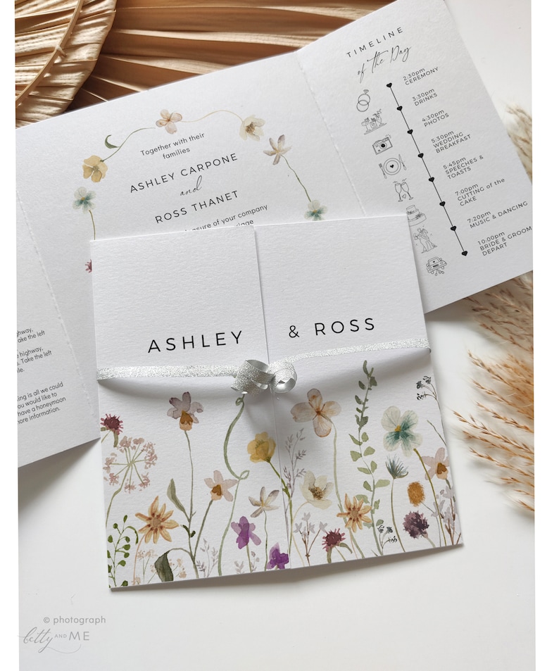 May include: A wedding invitation with a floral design. The invitation is folded in half and tied with a silver ribbon. The front of the invitation has the names Ashley and Ross written in black text. The back of the invitation has a timeline of the wedding day with icons and times.