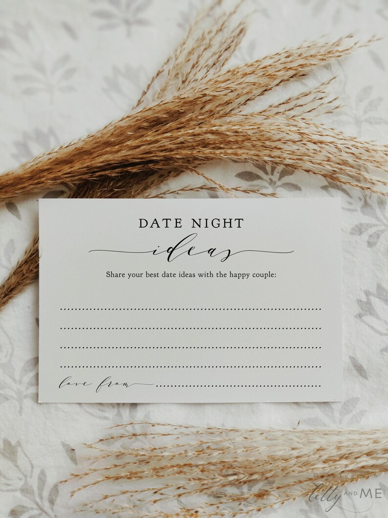 May include: A white card with the text "DATE NIGHT ideas" in black script. The card includes prompts to share date ideas and a space to write "love from." Dried, tan-colored ornamental grass is in the background.