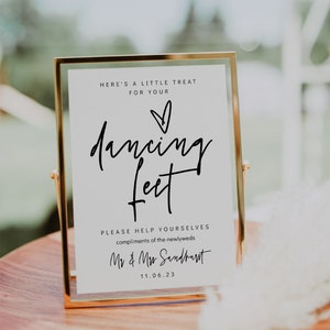 May include: A gold-framed sign with the words "dancing feet" in elegant script. The sign includes the text "Here's a little treat for your" and "Please help yourselves compliments of the newlyweds". The date 11.06.23 is also displayed.