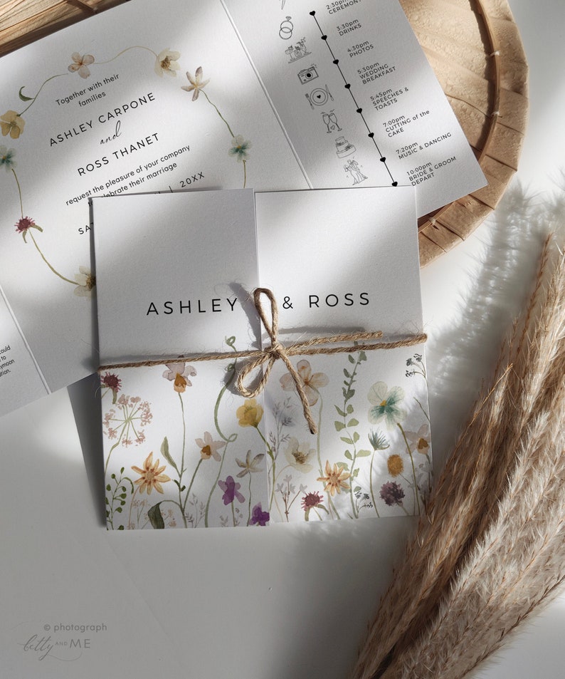 May include: A wedding invitation with a floral design. The invitation is folded in half and tied with twine. The front of the invitation has the names Ashley and Ross written in black text. The back of the invitation has a timeline of the wedding day.
