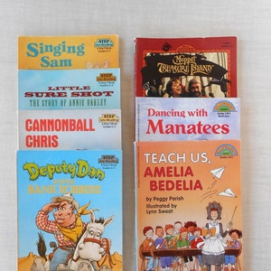 May include: A stack of colorful children's books with titles like "Singing Sam", "Little Sure Shot", "Cannonball Chris", "Deputy Dan and the Bank Robbers", "All Aboard Reading Muppet Treasure Island", "Dancing with Manatees", and "Teach Us, Amelia Bedelia".