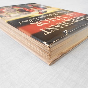 May include: A hardcover book titled "Merchant of Valor" by Lawrence Buddington Kelland. The book's cover features a red and black design with a partial illustration. The book's pages are aged, and the cover shows wear.