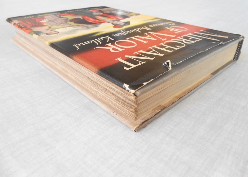 May include: A hardcover book titled "Merchant of Valor" by Lawrence Buddington Kelland. The book's cover features a red and black design with a partial illustration. The book's pages are aged, and the cover shows wear.