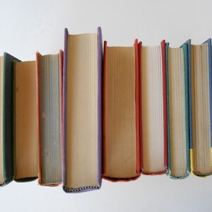 May include: A row of eight vintage hardcover books with different colored spines. The books are arranged in a row from left to right, with the spines facing the viewer. The books are all different sizes and have different titles and authors.
