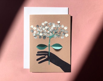 May include: A greeting card featuring a hand holding a paper flower with white petals and green leaves. The flower is made of paper and has a silver shimmer.