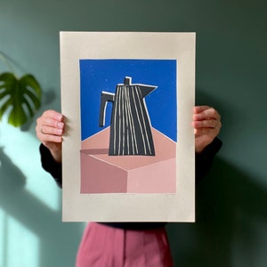 May include: A print of a black and white coffee pot with vertical lines, set against a blue sky and a pink geometric shape. The artwork is on a cream-colored paper.