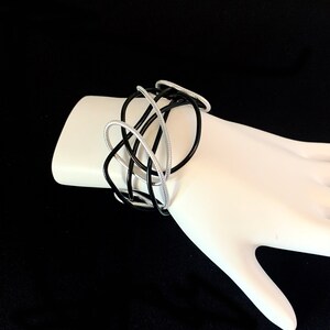 May include: A black and silver bracelet made of intertwined metal bands. The bracelet is designed in a modern, abstract style.