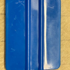 May include: A blue plastic squeegee with a smooth, flat surface and a rounded edge. The squeegee is used to apply and smooth out vinyl decals, stickers, and other adhesive materials.
