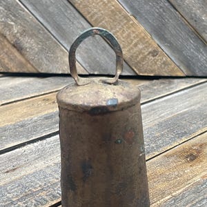 May include: A small, aged metal bell with a loop handle. The bell has a cylindrical body with a slightly flared base. The metal has a weathered, tarnished appearance with hints of green and red, suggesting age and exposure to the elements. The bell is likely used for decorative purposes.