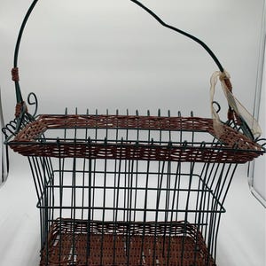 May include: A rectangular basket with a handle. The basket has a dark brown woven top and base, with a green metal frame. The handle is dark green with a brown woven grip. A sheer ribbon is tied to the side.