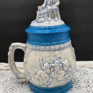 May include: A blue and white ceramic beer stein with a lid featuring a figurine of a man in traditional clothing. The stein has a handle and a detailed design of people in traditional clothing.