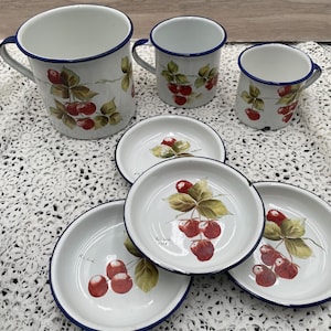 May include: Set of vintage enamelware kitchenware with a cherry motif. The set includes a large pitcher, two mugs, and four small plates. The plates and mugs have a blue rim and a white background with a hand-painted design of red cherries on green leaves.