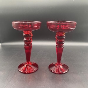 May include: Two ruby red glass candle holders. Each has a round base, a long stem with a series of rounded sections, and a shallow bowl-shaped top. The glass has a glossy finish. The candle holders are displayed against a dark background.