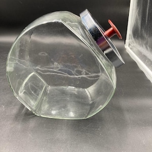 May include: A clear glass canister with a slanted opening and a metallic silver lid featuring a small, dark red knob handle. The container has a geometric, multi-faceted shape designed to sit at an angle, providing a wide access point.