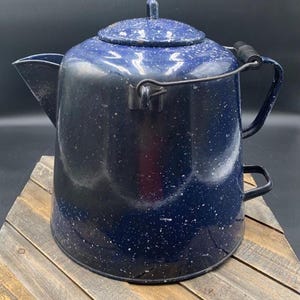 May include: A dark blue enamel coffee percolator with a white speckled pattern. The pot features a matching lid with a small handle, a wide pouring spout, a black metal wire bail handle with a wooden grip, and a small black side handle.