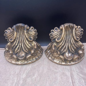 May include: A pair of matching wall brackets featuring a gold-toned finish with dark distressing. Each bracket displays a prominent fan-shaped acanthus leaf motif at the top, transitioning into a curved, semi-circular base with decorative molding.