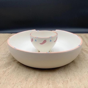May include: A cream-colored ceramic chip and dip serving bowl featuring a central integrated dip cup. The rim of the large bowl and the dip cup are decorated with a pattern of small pink chili peppers and light blue triangles on a white base.