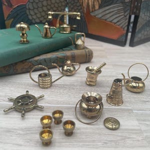 May include: A collection of miniature brass objects including a ship wheel, several small watering cans, a mortar and pestle, a small cauldron with a lid, tiny goblets, a lamp, and a scale, all arranged on a light-colored surface.