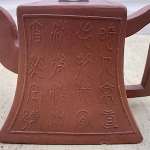 May include: A reddish-brown ceramic teapot with a square body and handle. The front of the teapot features an embossed rectangular panel with Chinese characters. The spout is curved, and the handle is rectangular.