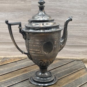 May include: An antique silver-plated teapot with a decorative handle and spout. The teapot features an ornate design with a detailed lid and a pedestal base. The silver finish gives it a vintage look, perfect for display or use.
