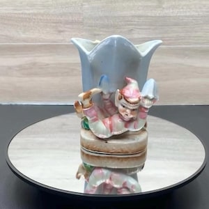 May include: A vintage ceramic vase with a light blue tulip-shaped design. A whimsical elf figurine in pink and white striped clothing is attached to the base. The elf is lying down with a mushroom in each hand. The vase is on a reflective surface.