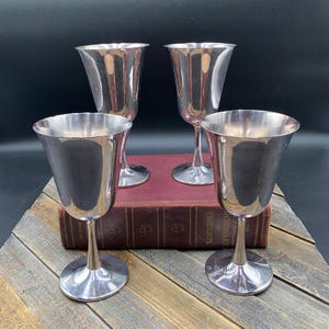 May include: A set of four silver-toned metal goblets with smooth, polished surfaces, flared rims, and slender stems resting on circular bases. The goblets feature a simple, unadorned design with a reflective finish that captures light across their curves.