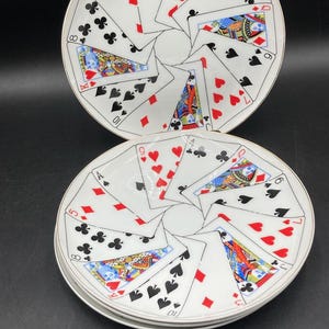 May include: A set of white ceramic plates featuring a circular arrangement of various playing cards, including hearts, diamonds, clubs, and spades, with face cards displaying colorful illustrations and gold-toned rims on each plate.