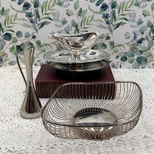 May include: A collection of silver serving pieces, including a gravy boat with a matching saucer, a decorative vase, and a wire basket. The items are arranged on a white lace tablecloth, with a floral wallpaper background.