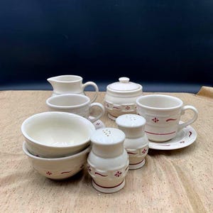 May include: A cream-colored ceramic dinnerware set featuring a red floral and line pattern. The collection includes a creamer, a lidded sugar bowl, two bowls, two cups with saucers, and a salt and pepper shaker set, all with a matching decorative motif.