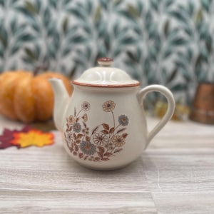May include: Cream-colored ceramic teapot with a floral design in brown, blue, and yellow. The teapot has a rounded body, a curved handle, and a lid with a small knob. The rim and lid are trimmed with a brown band. A pumpkin and fall leaves are in the background.