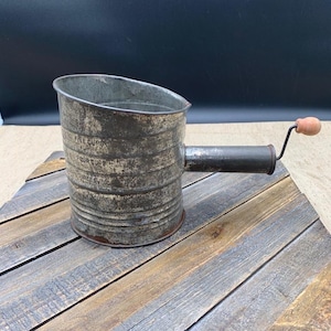 May include: A vintage metal flour sifter with a weathered gray finish and a cylindrical body. It features a side handle with a small wooden knob and printed measurement markings on the exterior that indicate levels up to five cups.