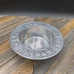 May include: A silver-colored, round ashtray with a recessed center. The rim is embossed with the number "100" and card suit symbols. The ashtray is made of metal and sits on a wooden surface.