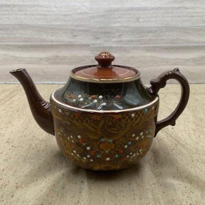 May include: A brown ceramic teapot with a dark green lid and handle. The teapot features a decorative pattern with gold, white, orange, and blue accents. The spout and handle are a darker shade of brown.