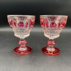 May include: Two vintage, ruby-colored crystal wine glasses. The glasses feature a clear bowl with ruby-colored circular accents and a ruby-colored stem and base. The glasses are displayed on a dark surface.