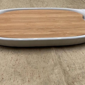 May include: An oval white ceramic serving tray with integrated side handles and a removable light brown wooden insert that fits snugly into the center, creating a dual-material surface for food presentation and serving.