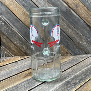 May include: A clear glass beer mug featuring the Oilers logo on both sides. The logo includes a white football helmet with red and blue accents. The mug has a handle and a wide, cylindrical shape.
