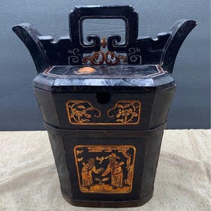 May include: Black wooden antique container with a handle and ornate gold detailing. The container features a rectangular shape with a lid and a lower section with a decorative panel depicting figures. The top has a handle and curved side elements.