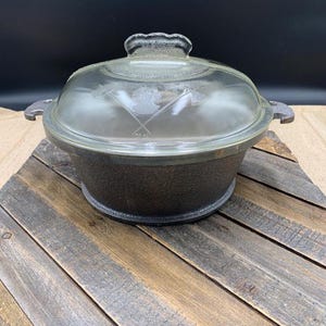May include: A round, dark brown cast iron Dutch oven with a textured exterior and two side handles. The pot features a clear, domed glass lid with a central handle and an etched graphic design showing a profile silhouette crossed by two axes.