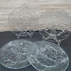 May include: Set of four clear glass dinner plates with a grapevine design. The plates are round and have a raised rim.