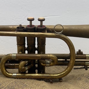 May include: Antique brass cornet with three valves and a flared bell. The instrument has a curved design with a mouthpiece and a finger ring. The cornet shows signs of age and wear, with a tarnished finish.
