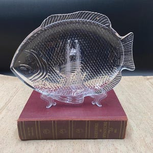 May include: A clear glass serving platter shaped like a fish with a textured scale pattern on the surface. The platter features a detailed head, fins, and tail, and is displayed on a clear plastic stand resting on a maroon hardcover book.